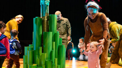 Scène du spectacle Arbre montrant des enfants et des interprètes autour d’une installation verte évoquant un arbre, sur une scène sombre.