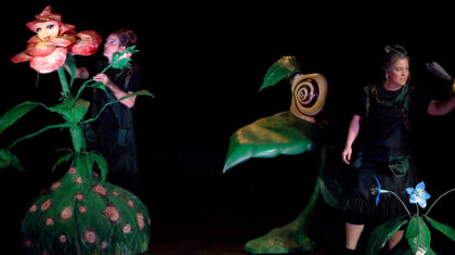 Scène du spectacle de marionnettes Un conte de jardin présentant des marionnettes végétales grandeur nature entourant deux manipulatrices sur une scène sombre.