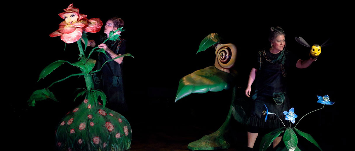 Scène du spectacle de marionnettes Un conte de jardin présentant des marionnettes végétales grandeur nature entourant deux manipulatrices sur une scène sombre.