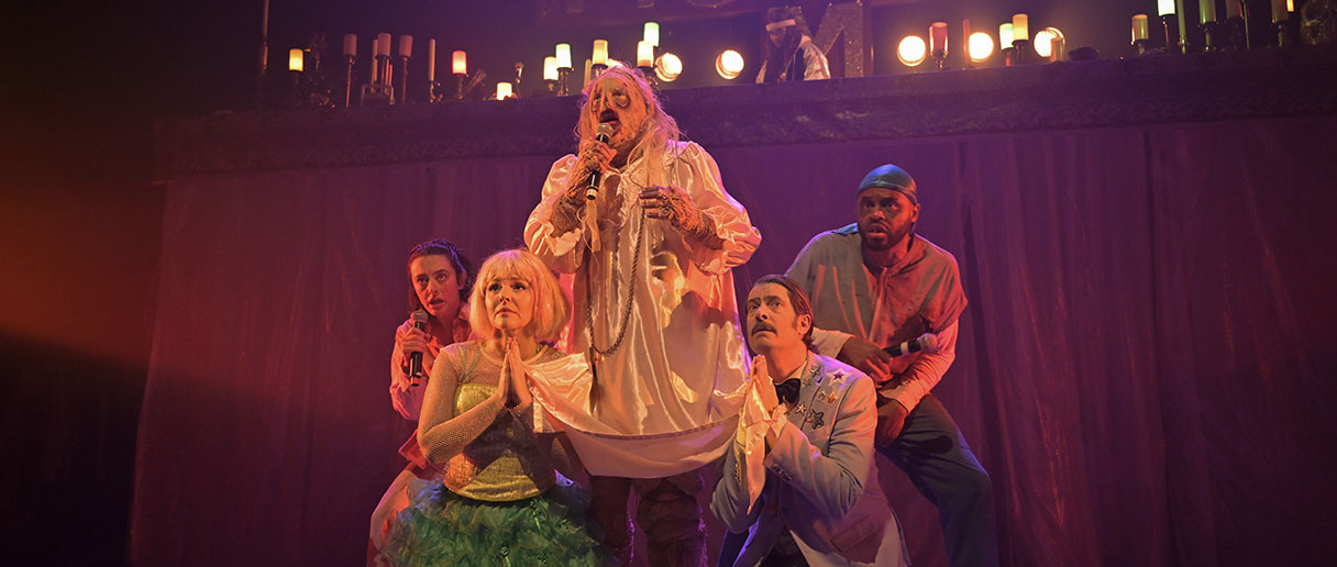 Une personne déguisée en zombie revêtant une robe blanche, dans la pièce de théâtre Mommy le retour, acompagnée de 4 comédiens/comédiennes dont deux qui font une prière et deux qui tiennent un micro.