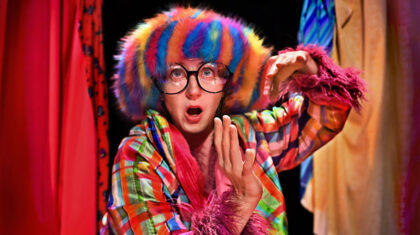 Spectacle Un nuage dans mon ventre — Sur scène, une interprète apparaît entre des pans de tissus colorés, parée d’un costume aux motifs vifs et d’une coiffe multicolore.