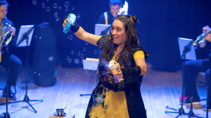 Spectacle La plume des périlleuses péripéties — Sur scène, une interprète manipule des objets ludiques pendant que des bulles flottent dans l’air, entourée de musiciens à vent, dans un univers théâtral et musical plein de fantaisie.