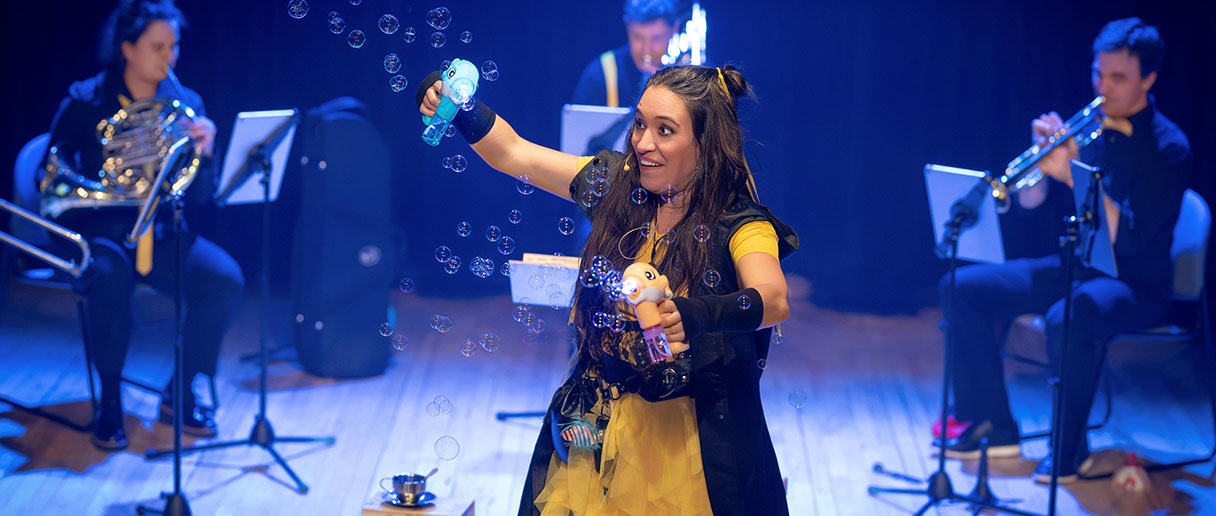 Spectacle La plume des périlleuses péripéties — Sur scène, une interprète manipule des objets ludiques pendant que des bulles flottent dans l’air, entourée de musiciens à vent, dans un univers théâtral et musical plein de fantaisie.