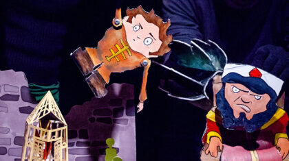 Spectacle Les routes ignorées — Théâtre d’objets et figures illustrées : un petit personnage en habit orange suspendu dans l’espace, tandis qu’un autre personnage, plus massif, apparaît à droite