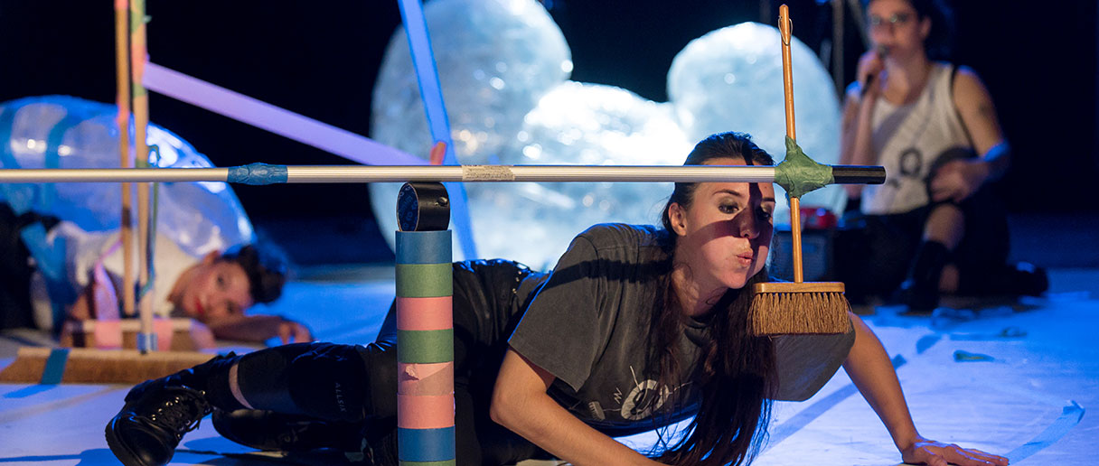 Théâtre : Scoooootch! — Sur scène, une interprète progresse au sol sous une structure bricolée de balais et de ruban adhésif, tandis que d’autres figures et matériaux envahissent l’espace.