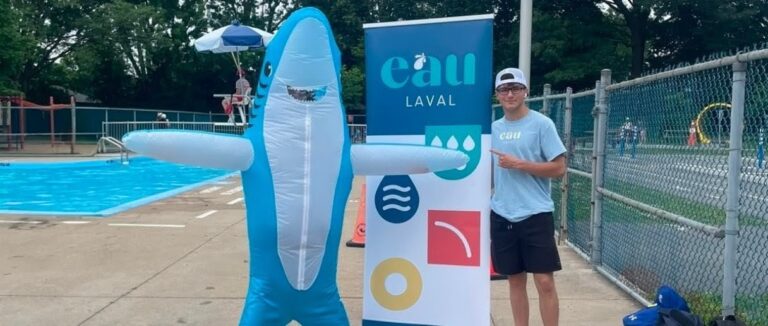 Une mascotte en forme de requin avec un jeune devant une pancarte de l'organisme Eau Laval